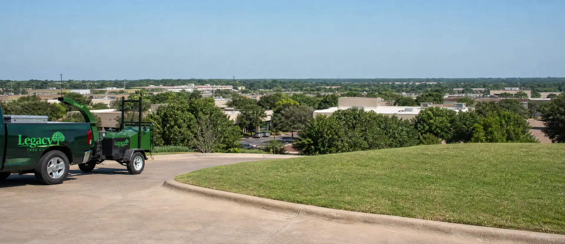 A Legacy Tree Care pickup and chipper parked on the access drive of a commercial property, with office buildings and mature tree canopy stretching to the horizon